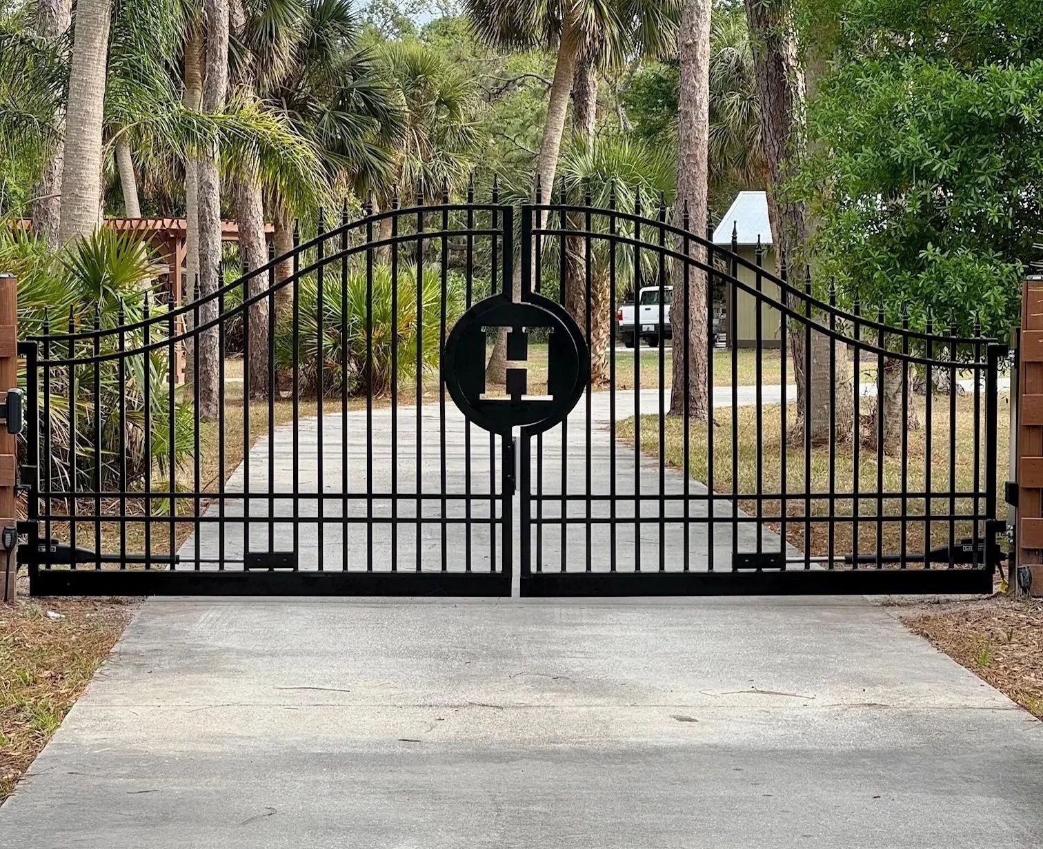 A black arched metal driveway gate with a central circle containing the letter H, set in a tree-lined residential area.
