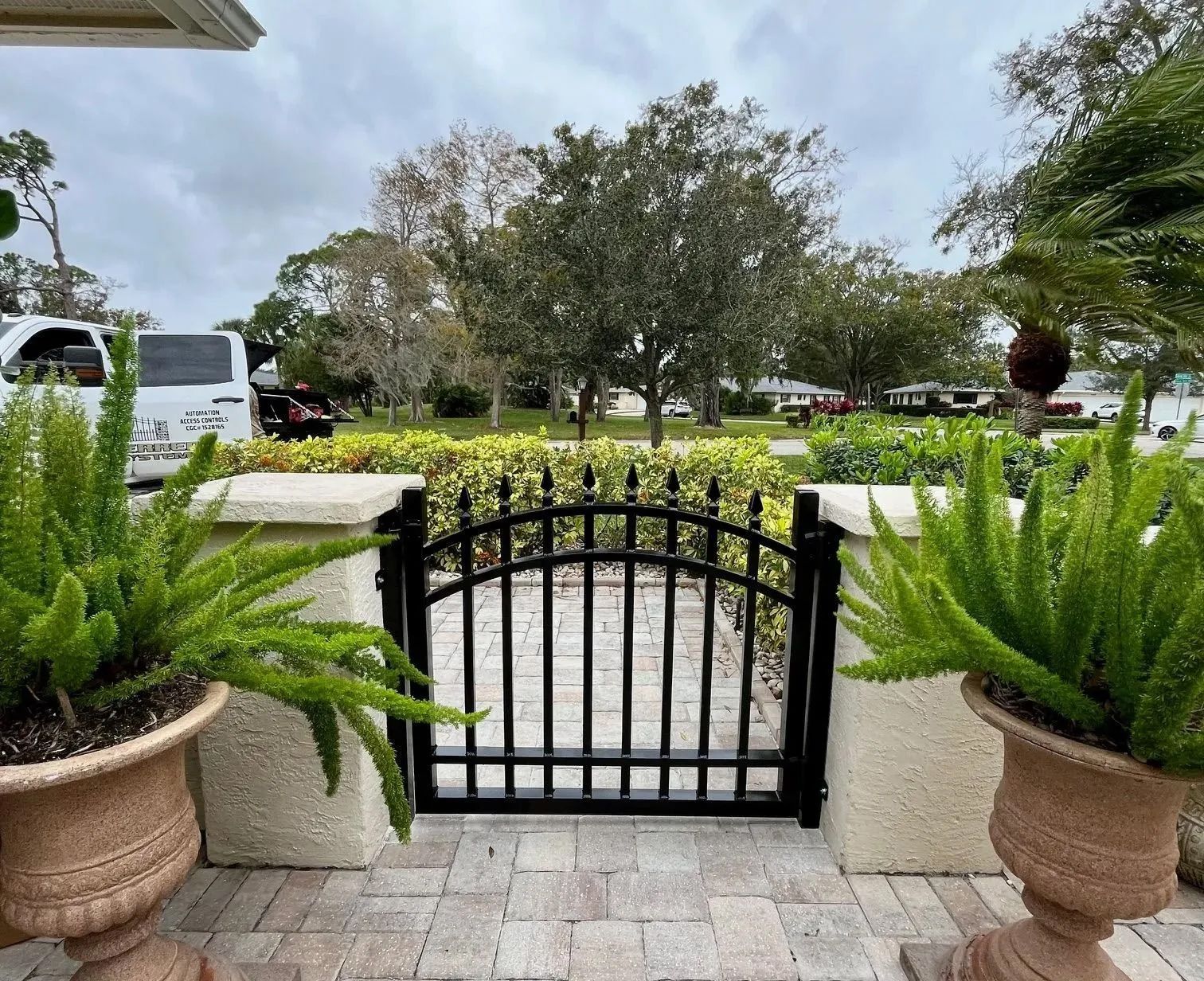 A black metal arched gate sits between two stone pillars, flanked by large potted ferns on a paved patio.