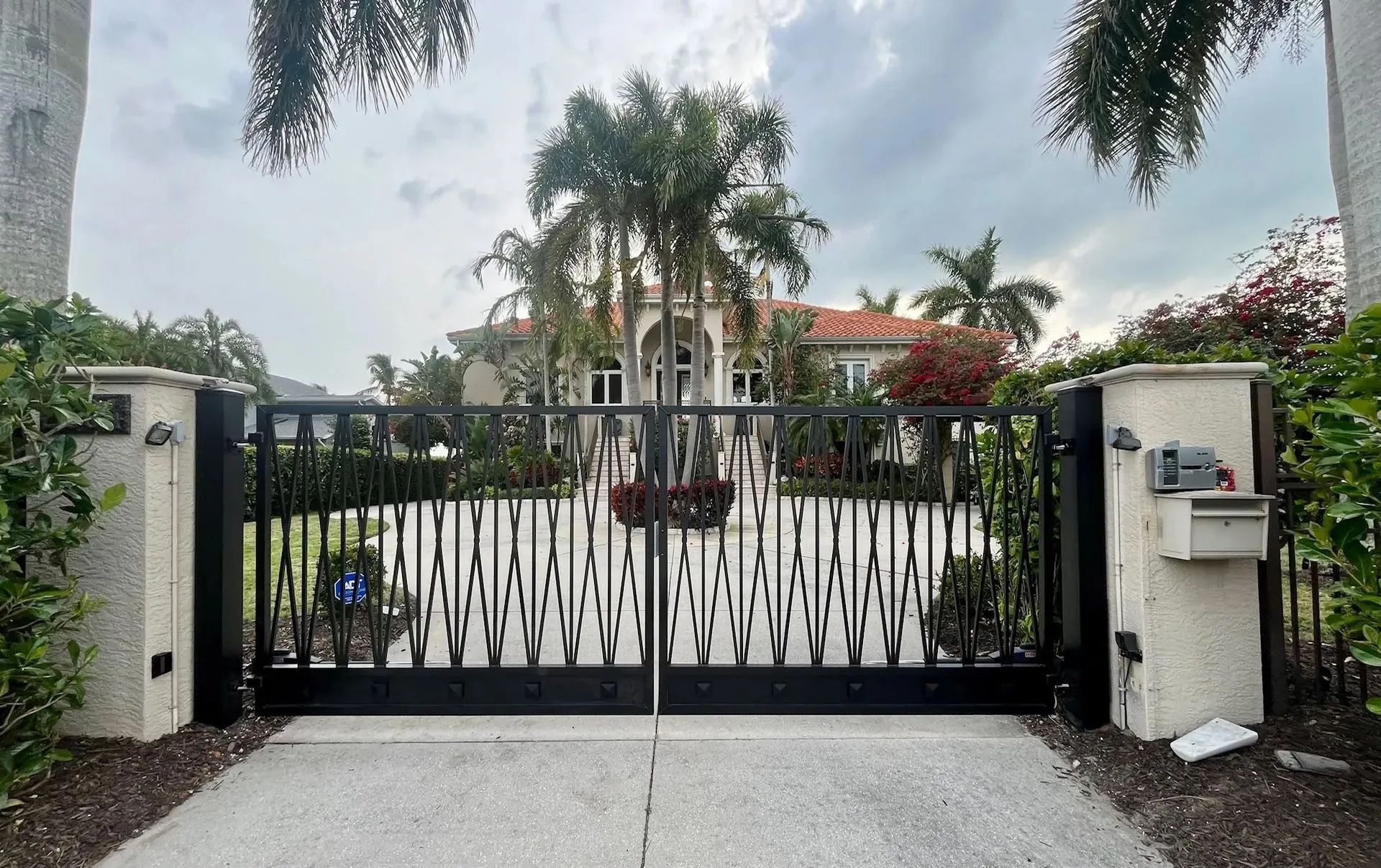 A black metal driveway gate stands in front of a white luxury house surrounded by palm trees and landscaping.