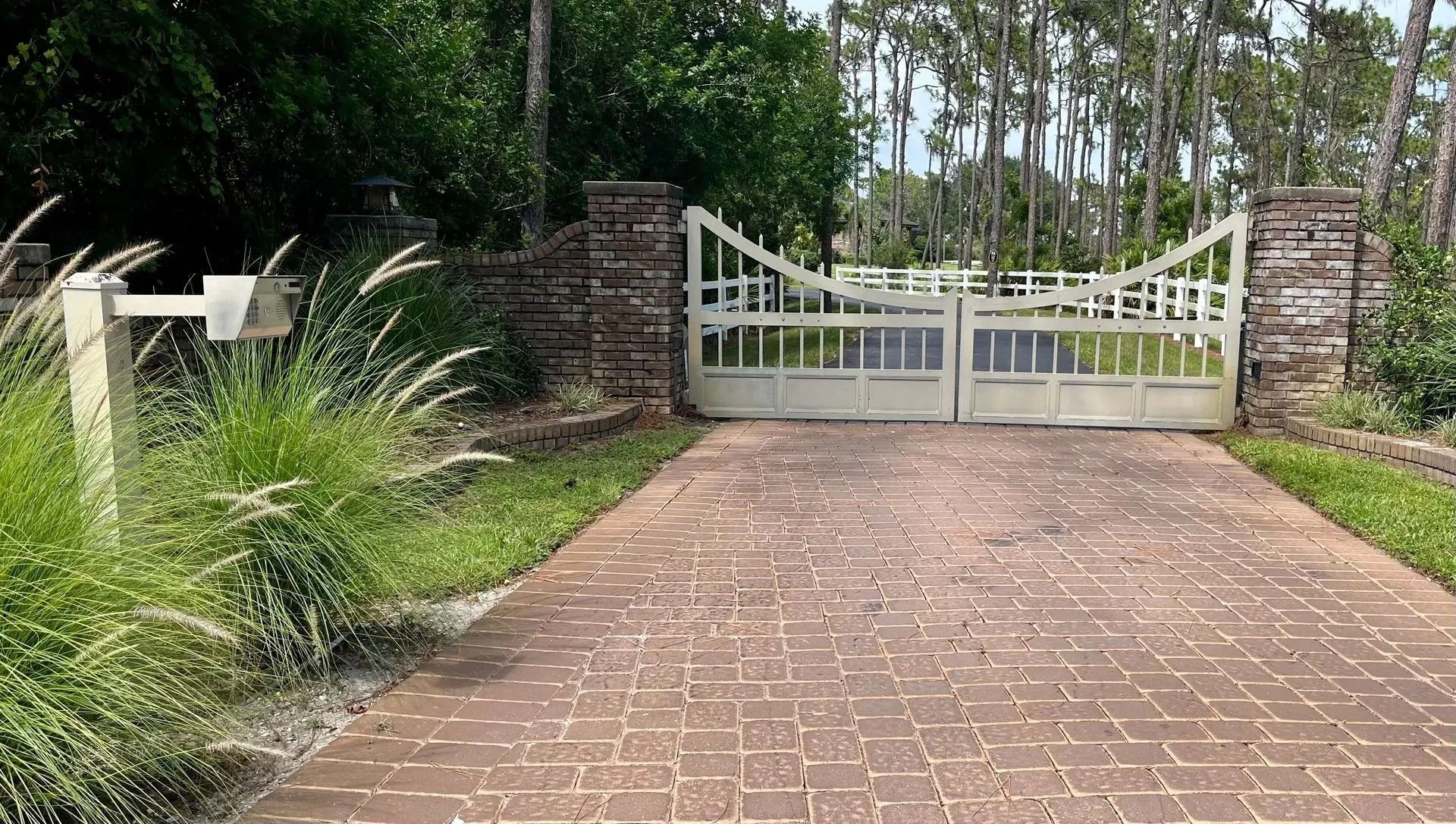 A brick driveway leads to white decorative gates flanked by brick pillars, surrounded by trees and tall grasses.