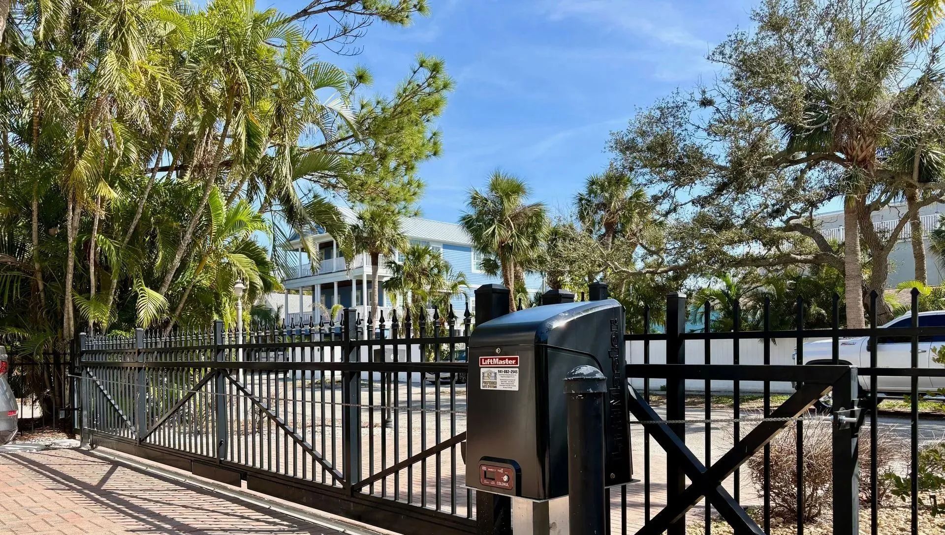 A black automated sliding gate operator mounted on a black metal fence in a sunny outdoor setting with trees and a house.