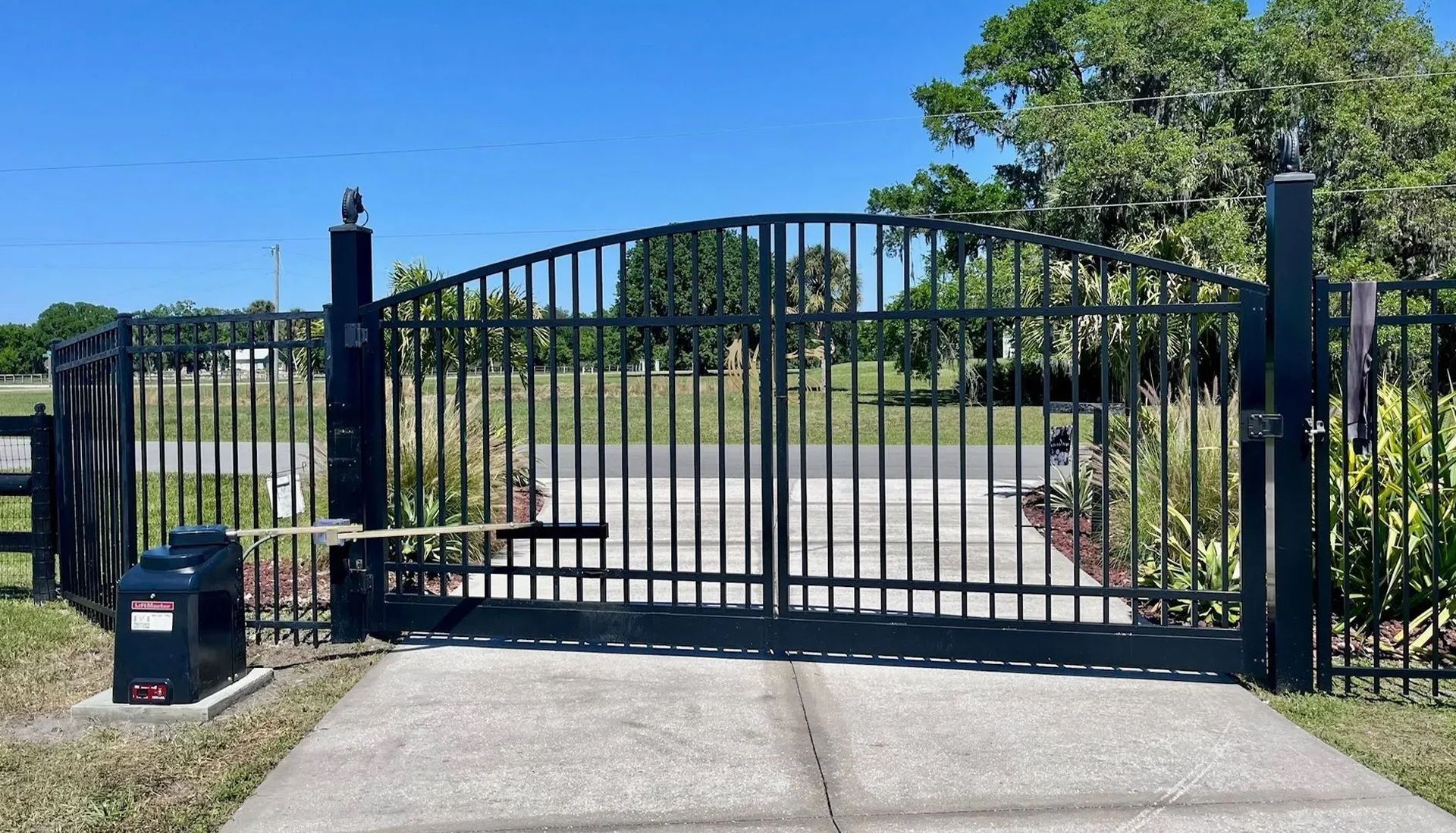 A black metal arched driveway gate with an automated operator box mounted on a concrete pad in a grassy, outdoor setting.