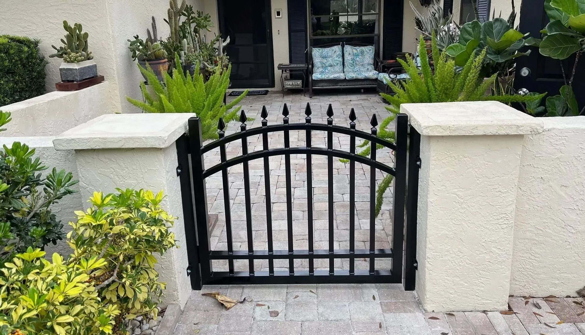 A black metal arched gate set between two white stucco pillars in front of a home entryway.