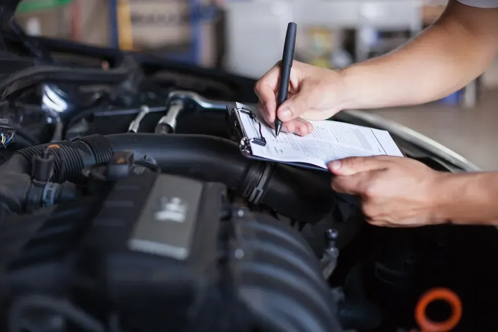 Mechanic inspecting a car engine, taking notes on a clipboard in a garage.