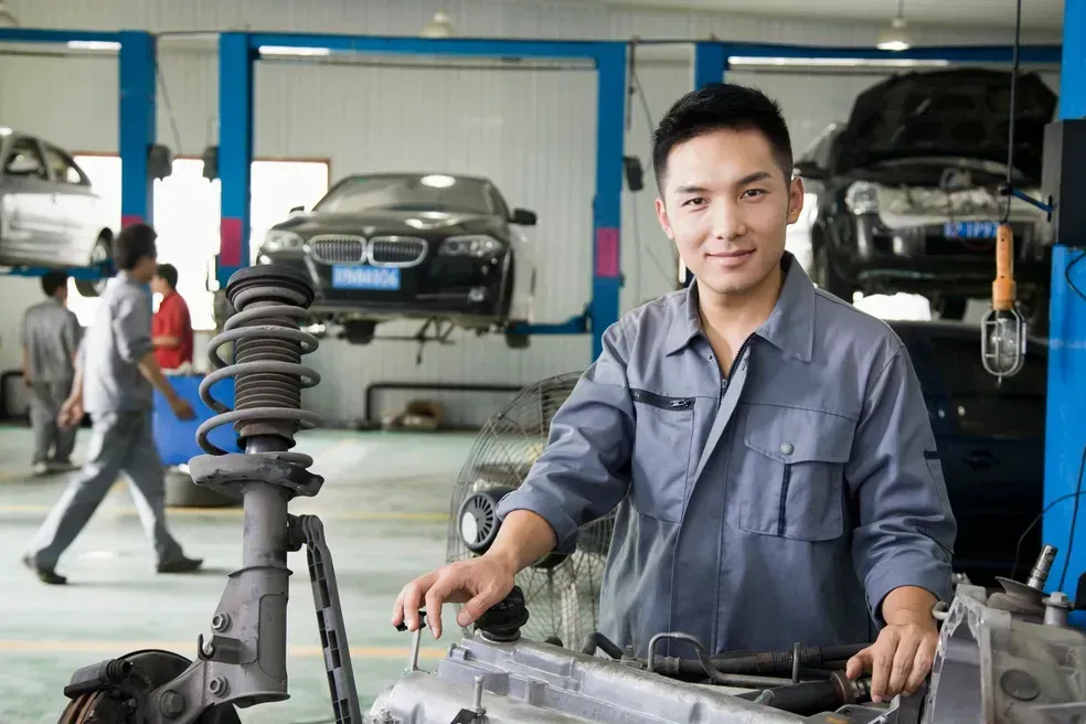 Mechanic in gray jumpsuit at a car repair shop, smiling, with cars being worked on in the background.