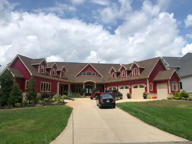 Red house with brown roof, dormers, and attached garage; cars in driveway, sunny day.