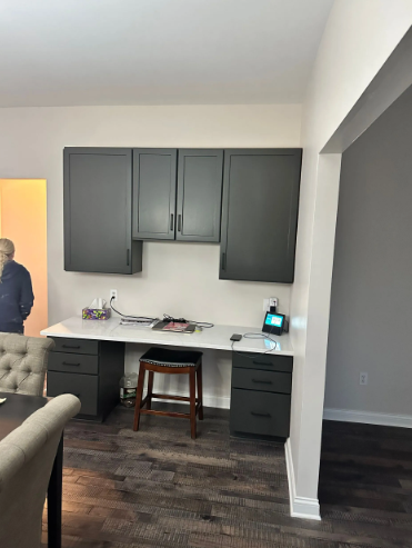 Built-in desk with dark gray cabinets above. A person stands left of the desk. The floors are a dark wood.
