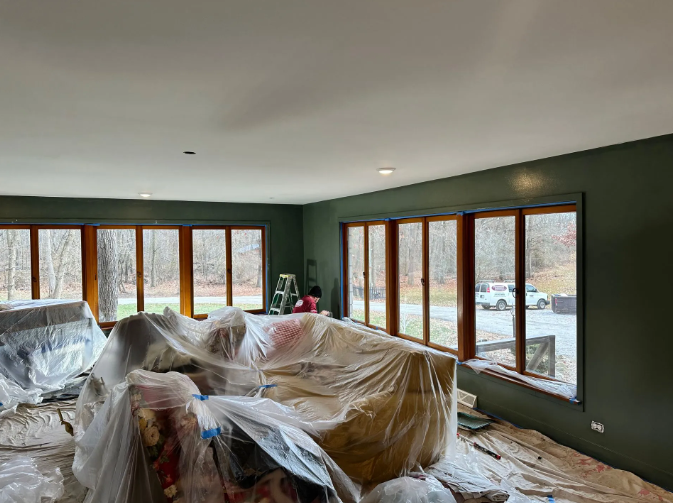 Room being painted; olive green walls, wood trim, white ceiling, furniture covered in plastic, view through windows.