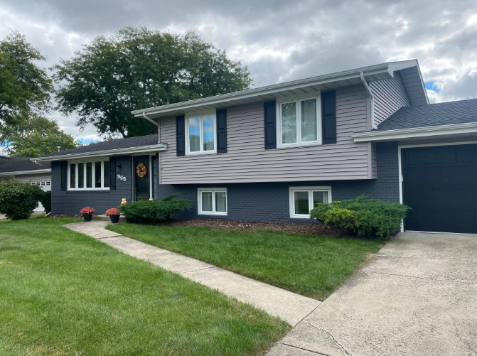 Gray and blue-painted house with black shutters, white-trimmed windows, and a paved walkway on a cloudy day.