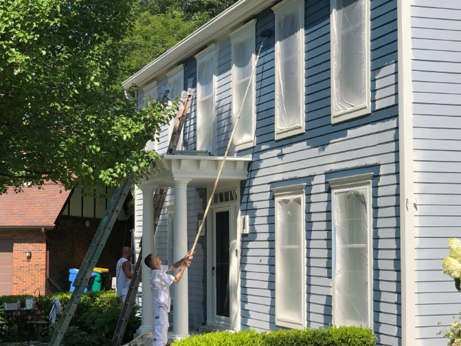 Two people painting a light blue house. One uses a long pole to paint near windows; the other is on a ladder.