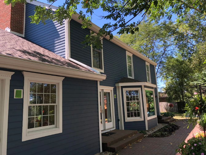 Blue two-story building with white trim and windows. Brick chimney on the left, foliage overhead.