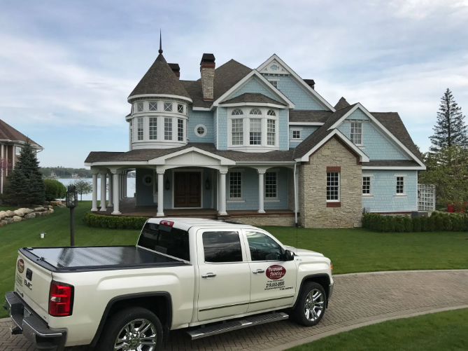 White pickup truck parked in front of a light blue Victorian house with a turret and covered porch, on a grassy lawn.