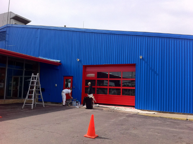 Blue building with a red garage door. Two people outside. One is painting, another is near the entrance. An orange cone is in the foreground.
