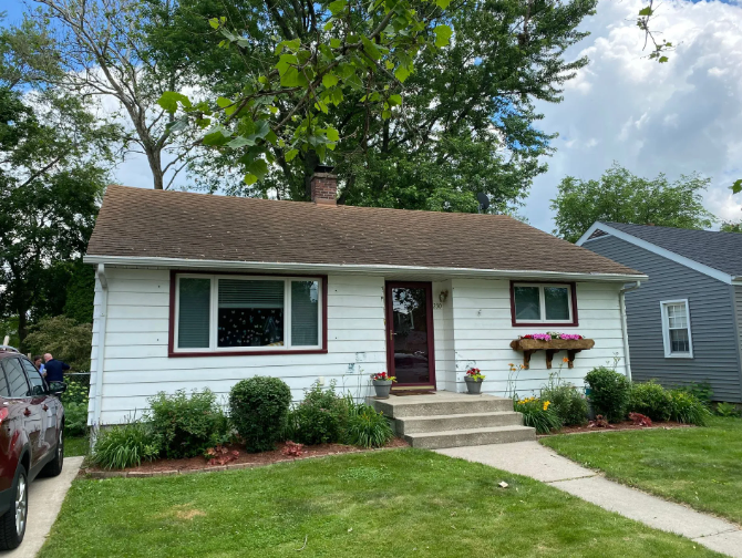 White house with brown roof, small porch, and green lawn. Flowers in window boxes and landscaping.