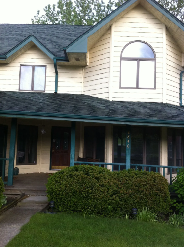 Two-story house with beige siding, dark green roof, and porch with blue railing. Front yard with green grass.