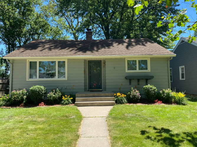 Green house with a brown roof and a concrete walkway. Landscaping with shrubs and flowers.