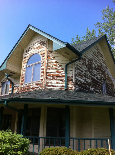 House with severely peeling paint on siding and trim, blue sky background.