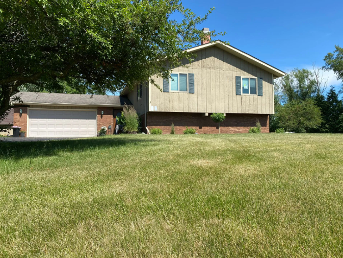 House with attached garage, beige siding, brick base, and large grassy yard under a bright blue sky.