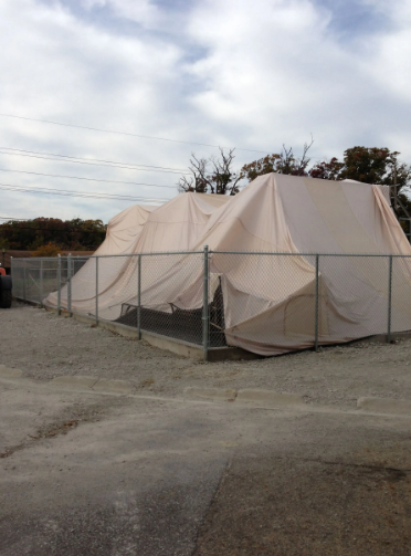 Fenced outdoor structure covered in draped light-colored fabric, gravel ground, overcast sky.