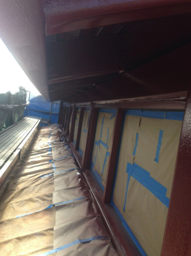 Brown painted eaves and window frames, blue tape, paper covering, outdoors.