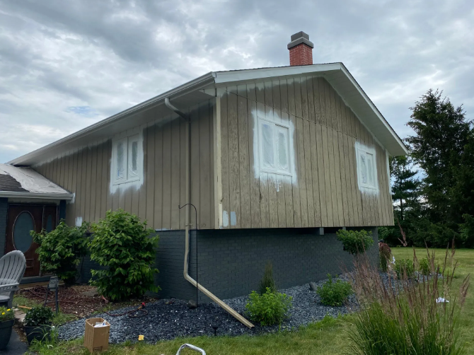 House exterior with white-trimmed windows and partially painted tan siding, under a cloudy sky.