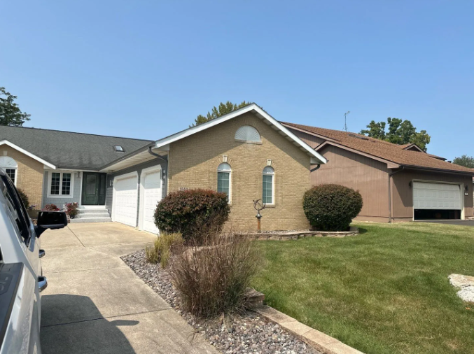 House with stucco exterior, attached garages, and a driveway on a sunny day. Green lawn with bushes.