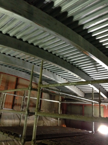 Steel beams and corrugated metal ceiling of a curved roof structure under construction, scaffolding in foreground.
