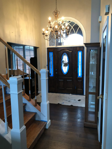 Entryway with a dark wood door and arched window. Chandelier hangs above, with staircase on the left and cabinet on the right.