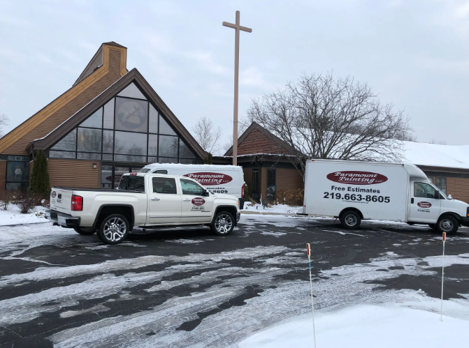 Two white service vehicles parked outside a church on a snowy day. A cross is visible.