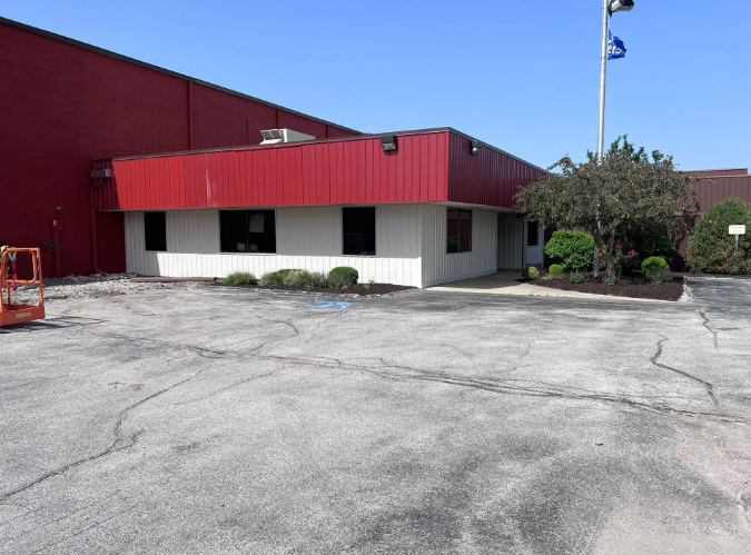 Red and white industrial building with cracked asphalt parking lot, blue sky, and a few bushes.