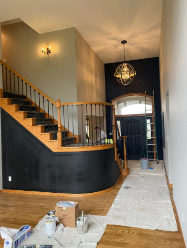 Entryway with staircase and black accent wall, being painted. Hardwood floors, chandelier, and a ladder are visible.