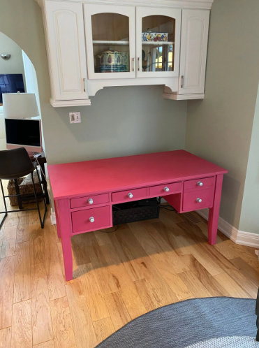 Pink desk with drawers, under white cabinets, in a room with hardwood floors and green walls.