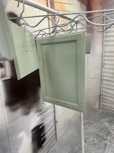 Cabinet doors, newly painted light green, hanging on metal racks in a spray booth.