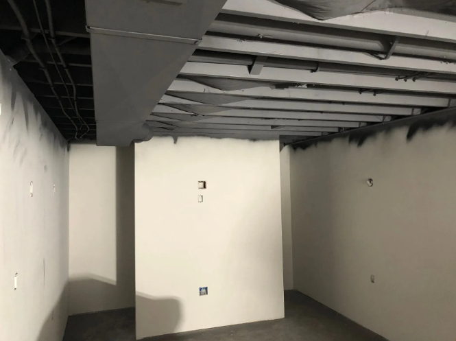 Empty unfinished room, white walls, gray floor, exposed ceiling beams, electrical outlets.