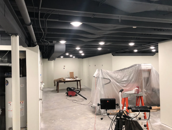 Basement under construction, with exposed beams and recessed lighting. White walls, gray floor, tools present.