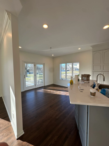 Interior of a room with dark wood floors, white walls, and a kitchen counter. Sunlight streams in.