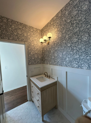 Bathroom with floral wallpaper, white wainscoting, wooden vanity with gold faucet, and globe light fixture.