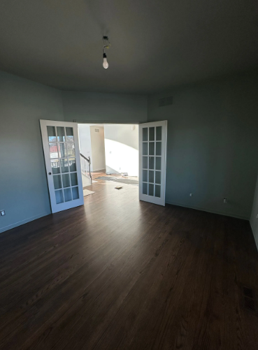 Room with dark wood floors, blue-green walls, white French doors, and a distant doorway with sunlight.