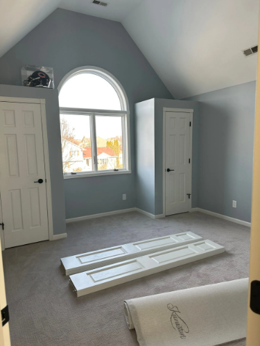 Bedroom with pale blue walls, arched window, two white doors, and light gray carpet.