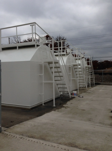 White fuel storage tanks with attached ladders and a platform, set against a cloudy sky.