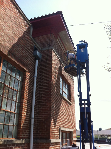 Workers on lift repairing brick building's roof; blue lift, red roof, overcast sky.