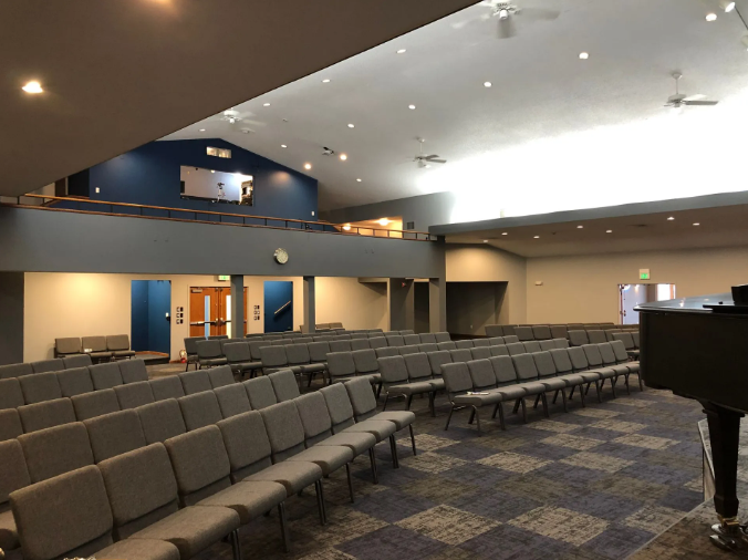 Interior of a church sanctuary with rows of gray seating, a stage area with a grand piano, and an upper level.