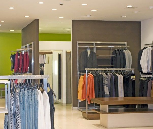Clothing racks with hanging garments and a wood display table inside a retail store with green and brown wall accents.