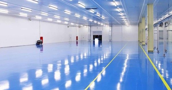 A large, empty industrial room with a glossy blue epoxy floor, white walls, and rows of bright ceiling lights.