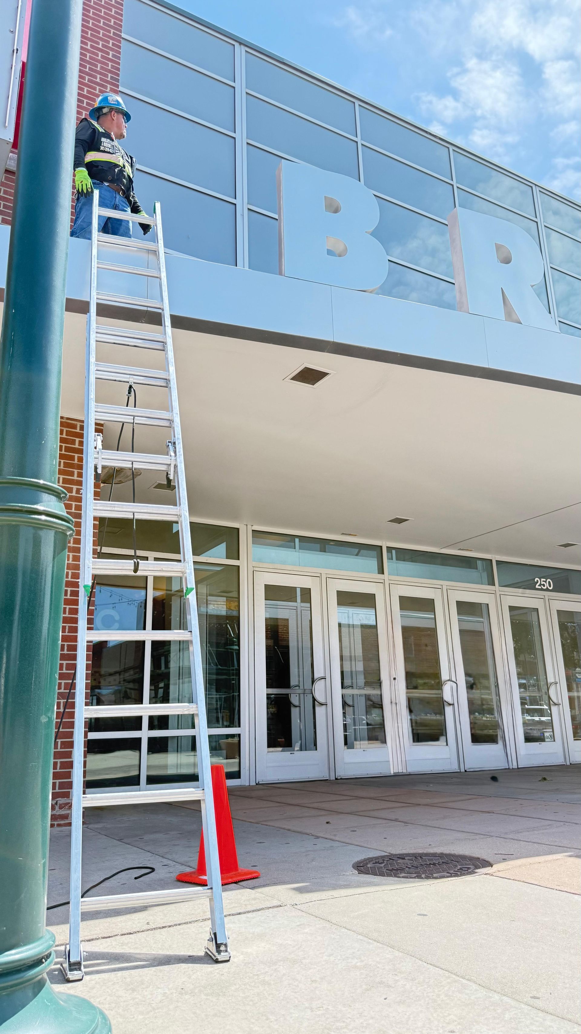Person on a ladder working on the letters above an entrance. A red cone and a lamppost are visible.