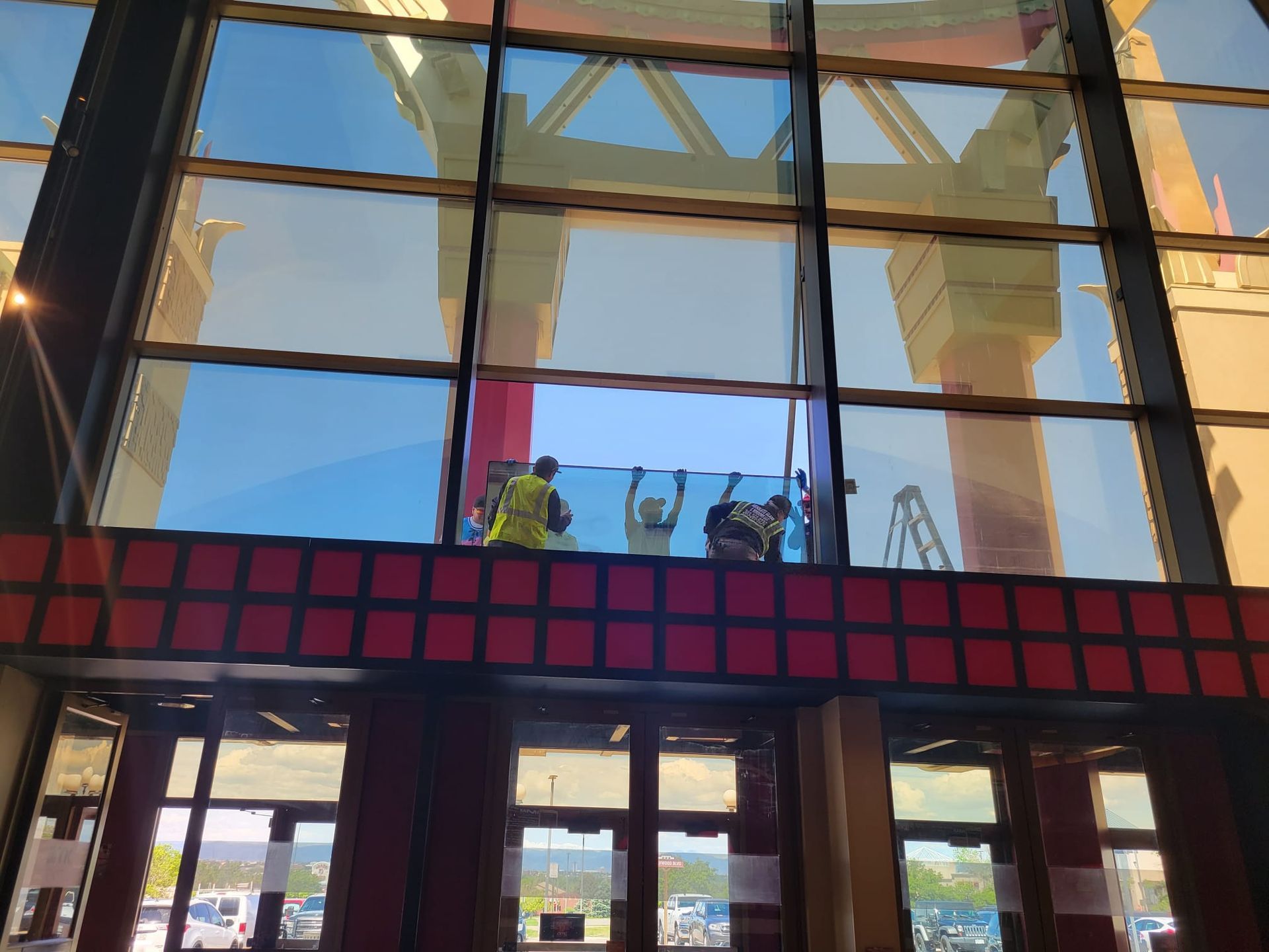Workers installing large window pane at building entrance. One in a yellow vest.