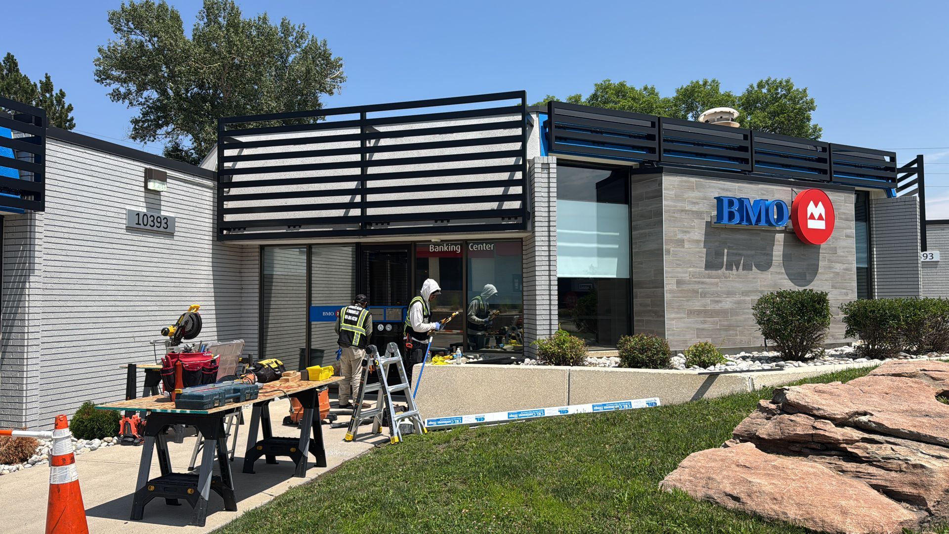 Construction at a Bank, workers near front entrance, tools visible. Blue and red bank logo. Sunny day.