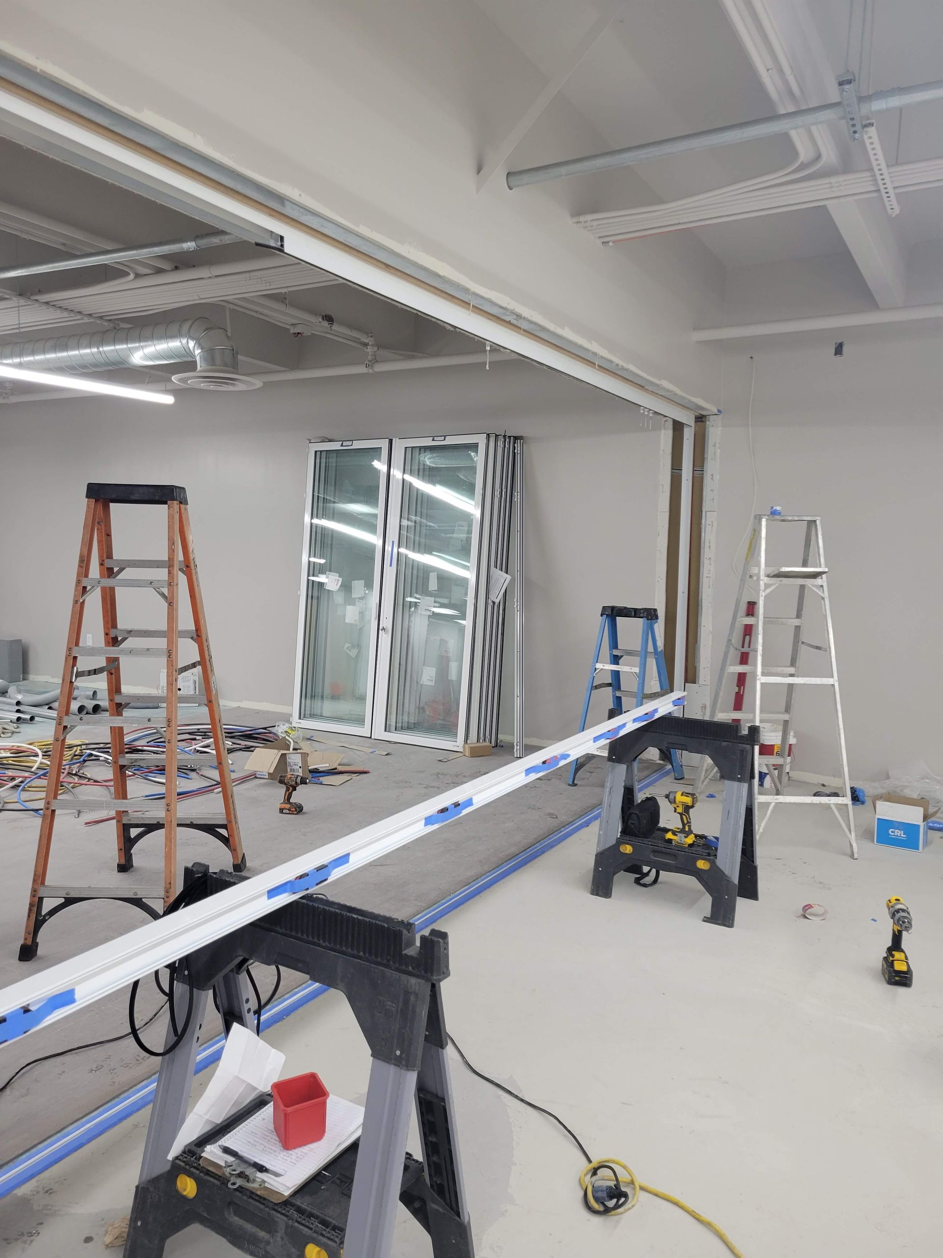 Construction scene: Workers install windows. Ladders, saw, and materials are in a white-walled room with bright overhead lighting.