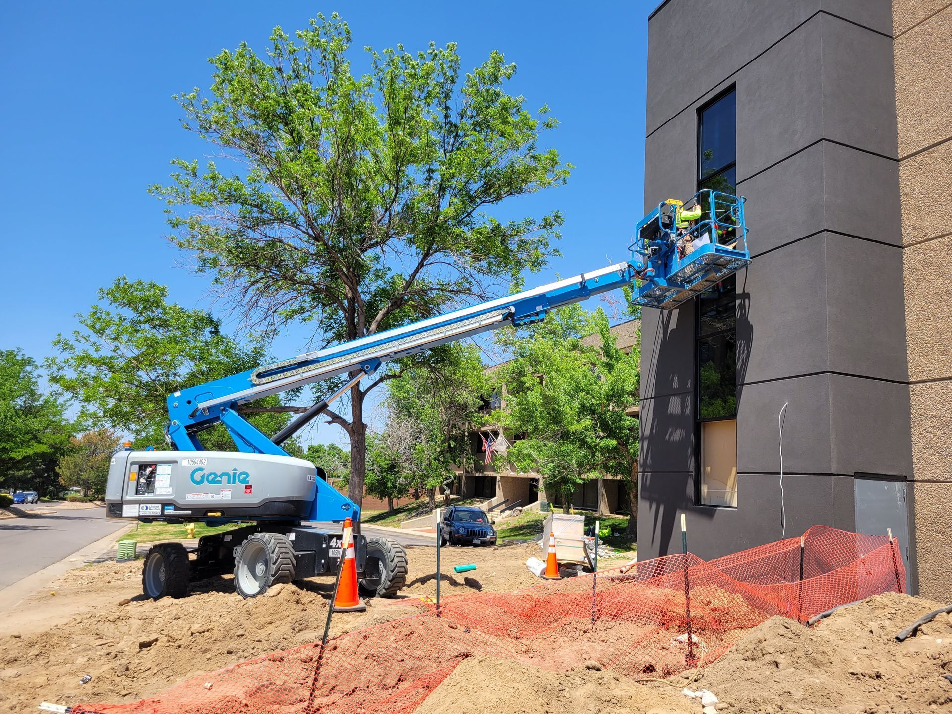 Construction worker in a lift repairing building exterior. Blue lift, red fencing, and brown building. Sunny day.