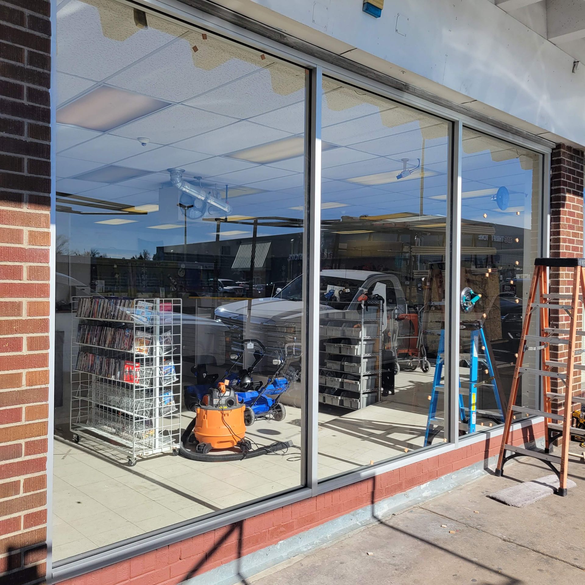 Exterior view of a store's large window. Interior shows construction, ladders, and displays. Brick and metal frame.